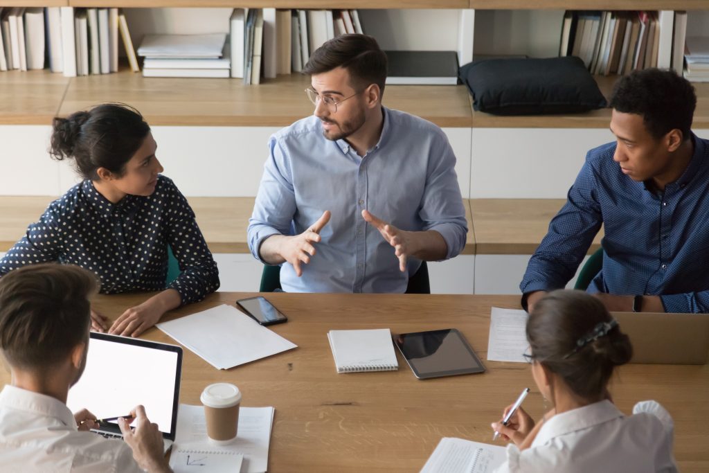 Five people sit around a table having a discussion. Notebooks, tablets, a laptop, and coffee are on the table. A man in the center is speaking about best practices, while others listen and take notes, fostering team building.