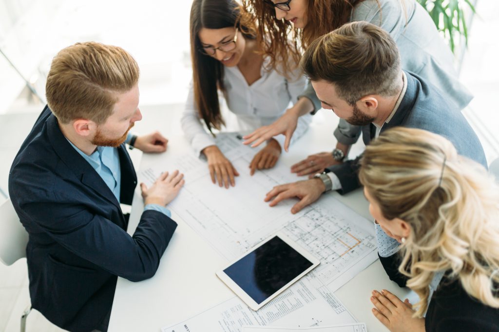 A group of people discussing architectural blueprints around a table with a tablet, focusing on innovative breakthroughs.